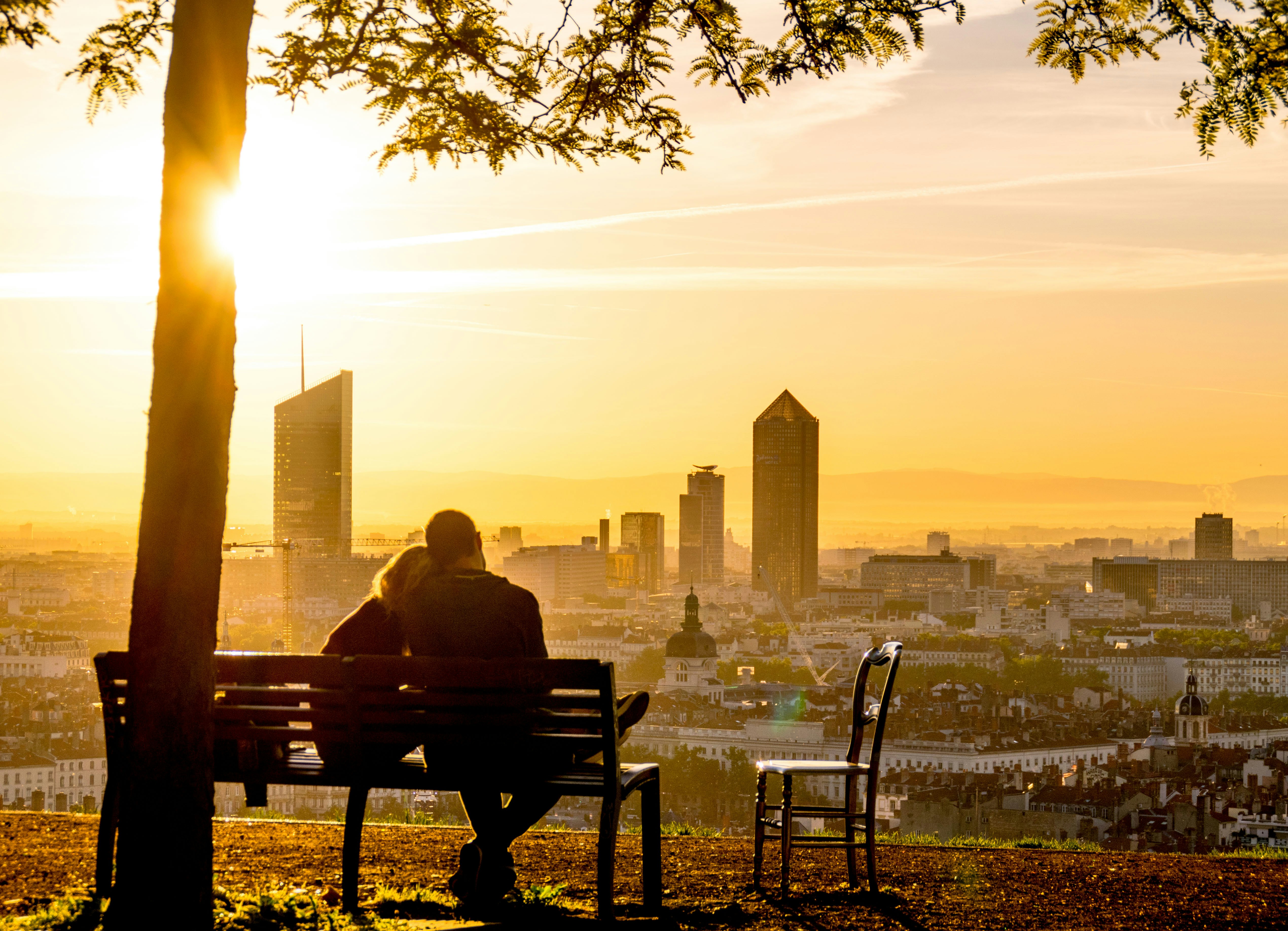 A couple sitting on a park bench while looking down at a city in France.